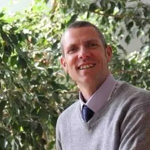 man in shirt and tie with light grey sweater smiling in front of a lot of greenery